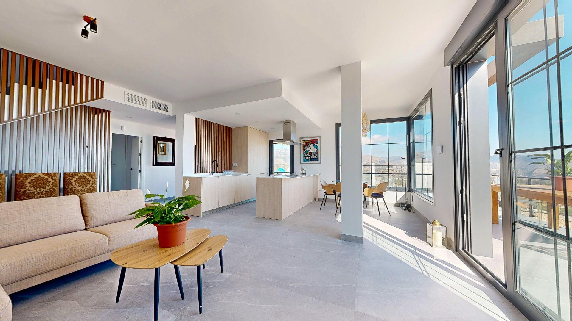 Open-plan living area with a beige sofa, wooden coffee tables, a kitchen island, and large windows with a mountain view outsideby the balcony.