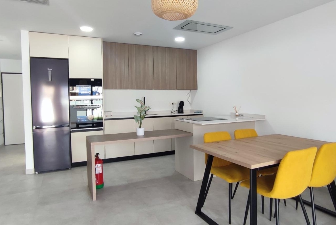 Open-plan kitchen and dining area with a stainless fridge, beige cabinets, and a wooden dining table with yellow chairs.