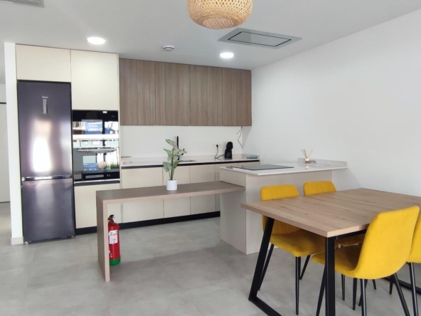 Open-plan kitchen and dining area with a stainless fridge, beige cabinets, and a wooden dining table with yellow chairs.