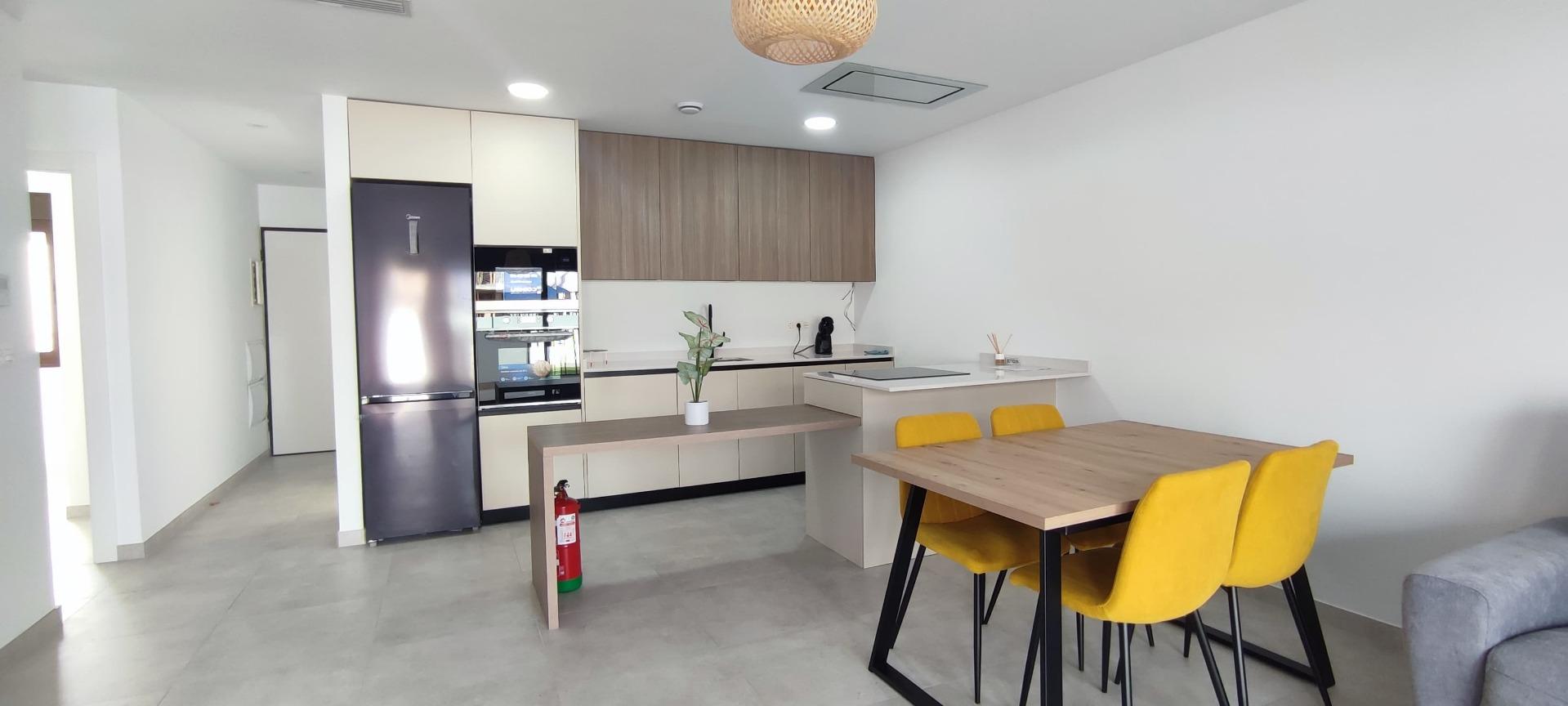 Open-plan kitchen and dining area with a stainless fridge, beige cabinets, and a wooden dining table with yellow chairs.