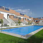 Modern apartment complex with tan and white facades, glass balconies, and a rectangular blue pool in the foreground under a clear blue sky.