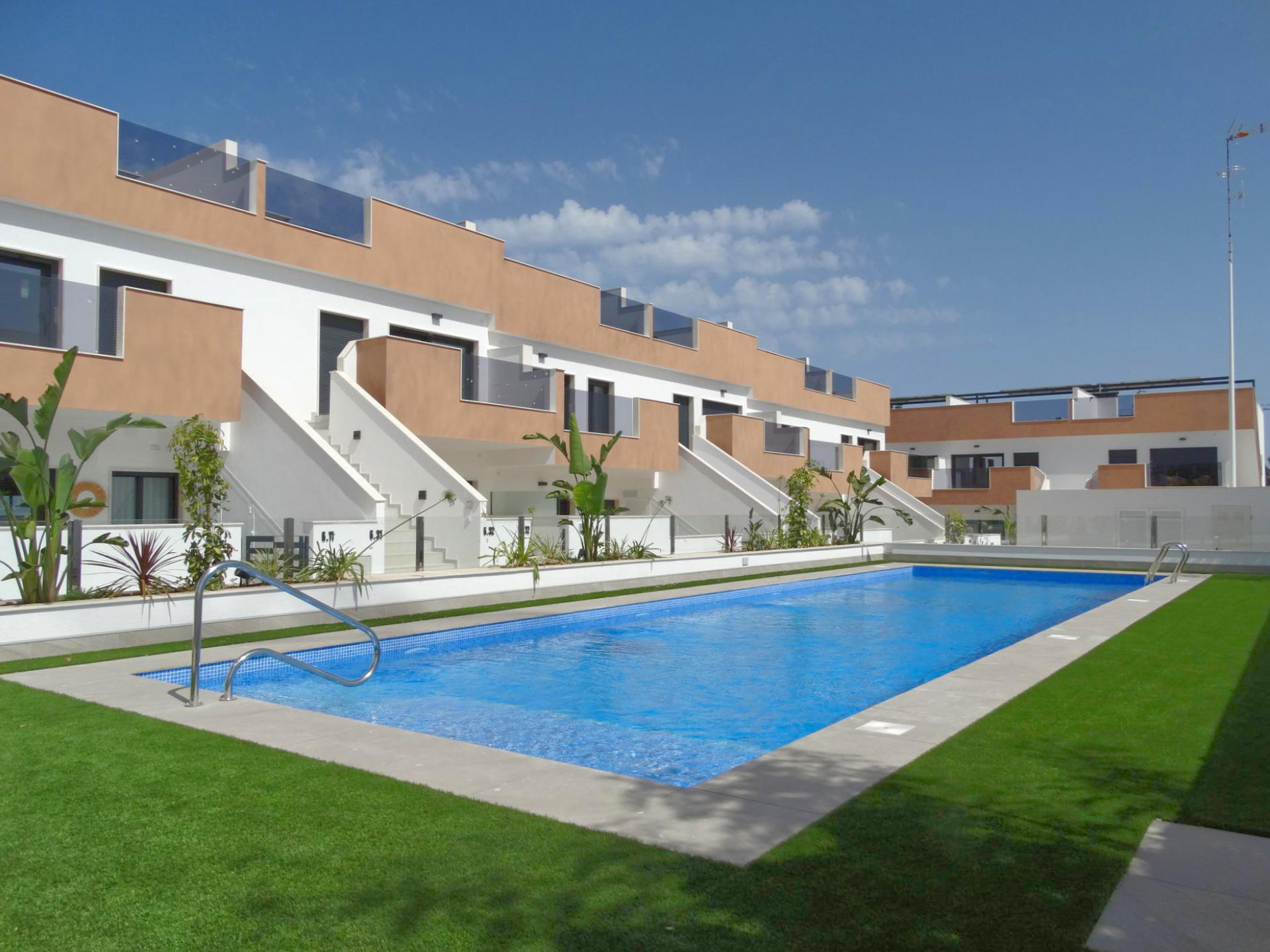 Modern apartment complex with tan and white facades, glass balconies, and a rectangular blue pool in the foreground under a clear blue sky.