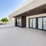 Open terrace of a modern building with large glass sliding doors, white and charcoal walls, and a wide gray-tiled floor under a recessed overhang, blue sky above.