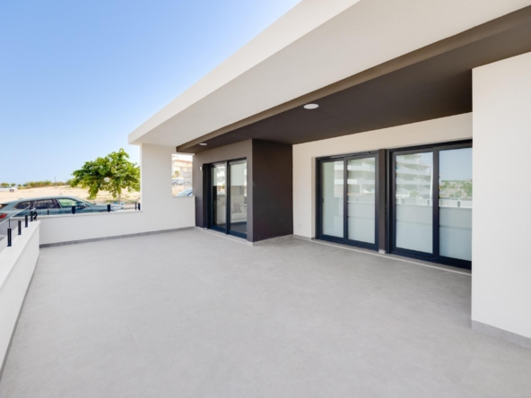 Open terrace of a modern building with large glass sliding doors, white and charcoal walls, and a wide gray-tiled floor under a recessed overhang, blue sky above.