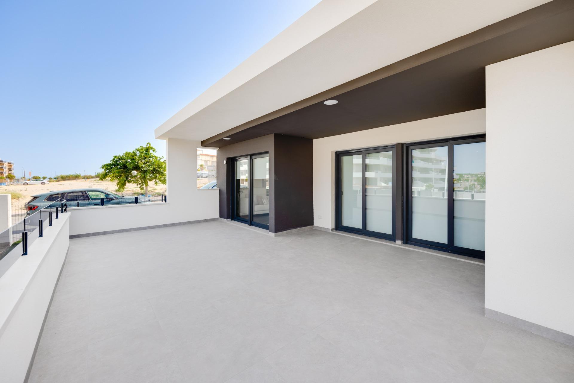 Open terrace of a modern building with large glass sliding doors, white and charcoal walls, and a wide gray-tiled floor under a recessed overhang, blue sky above.