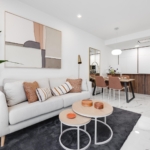 Open-concept living area with a white sofa, orange cushions, a tall green plant, and abstract wall art near a dining table and modern kitchen.