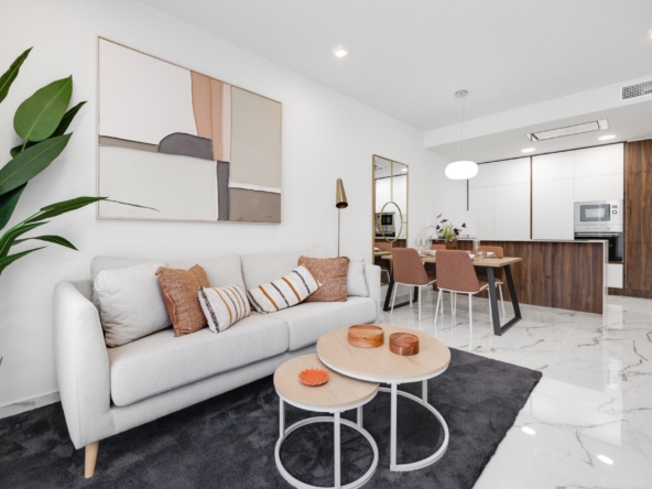 Open-concept living area with a white sofa, orange cushions, a tall green plant, and abstract wall art near a dining table and modern kitchen.