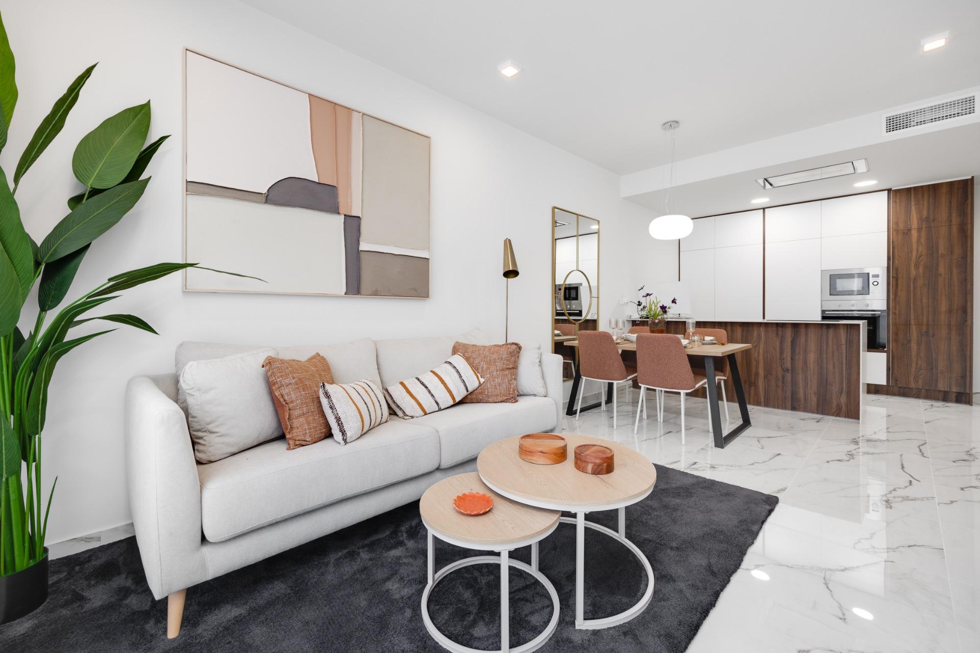 Open-concept living area with a white sofa, orange cushions, a tall green plant, and abstract wall art near a dining table and modern kitchen.