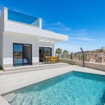 Modern white villa with glass balcony, outdoor dining table, and a rectangular pool under a clear blue sky, palm trees nearby.