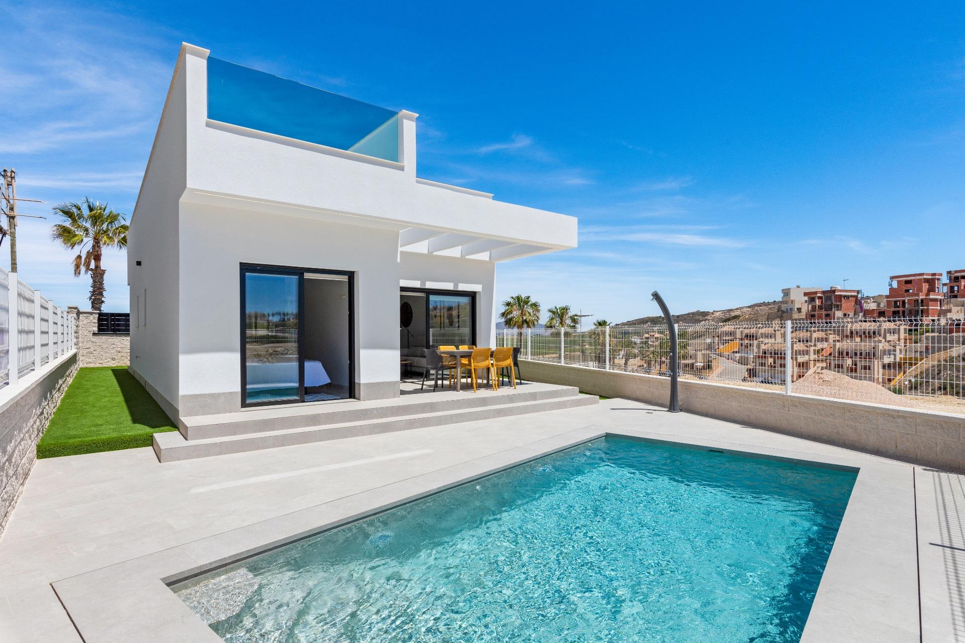 Modern white villa with glass balcony, outdoor dining table, and a rectangular pool under a clear blue sky, palm trees nearby.