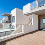 Contemporary apartment terrace with glass railings, wooden-tiled deck, and a sliding glass door opening to a living room under a blue sky.