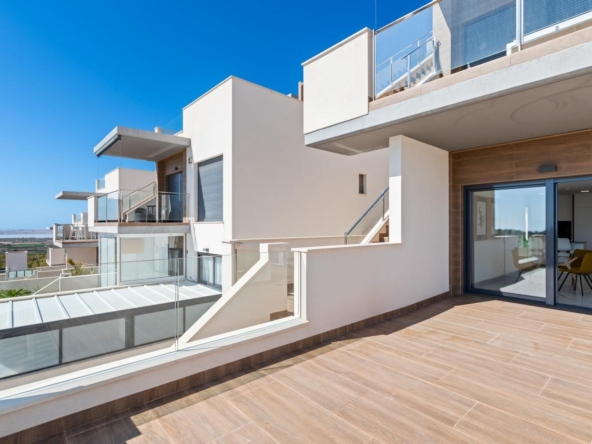 Contemporary apartment terrace with glass railings, wooden-tiled deck, and a sliding glass door opening to a living room under a blue sky.