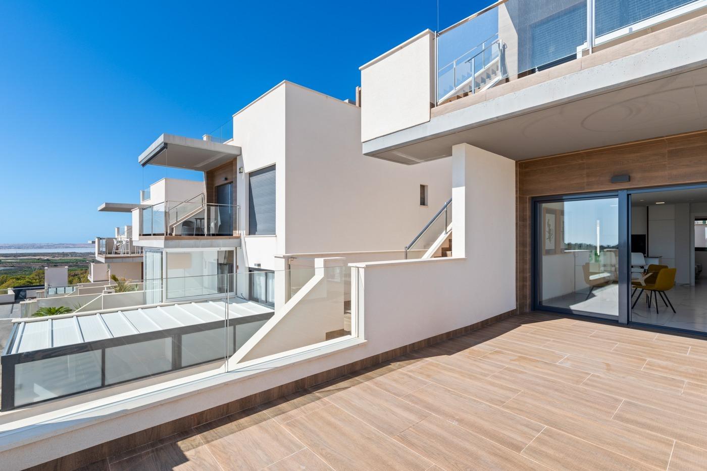 Contemporary apartment terrace with glass railings, wooden-tiled deck, and a sliding glass door opening to a living room under a blue sky.