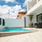 Modern backyard patio with a rectangular pool, beige stone deck, and a black metal dining set beside glass doors.