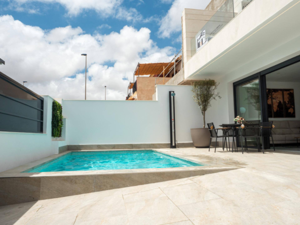 Modern backyard patio with a rectangular pool, beige stone deck, and a black metal dining set beside glass doors.