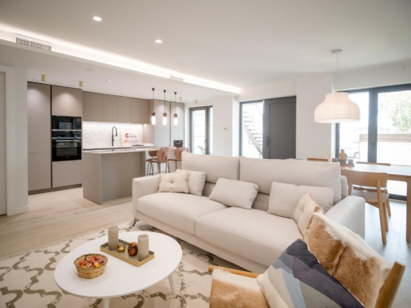 Open-plan living area with a light-gray kitchen, white sofas, and a dining table by large glass doors letting in natural light.