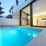 Modern white house with large glass doors opening to a warmly lit interior, beside a turquoise pool at dusk.