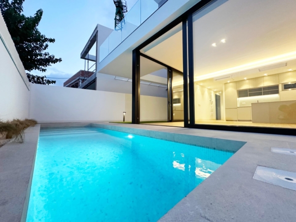 Modern white house with large glass doors opening to a warmly lit interior, beside a turquoise pool at dusk.