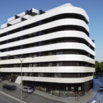 Futuristic white building with rounded, stacked balconies along a street; cars and pedestrians nearby.