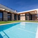 Modern villa with large glass doors, wooden beams, and a rectangular blue-tiled pool in the yard. Suitable for a luxury home exterior image.