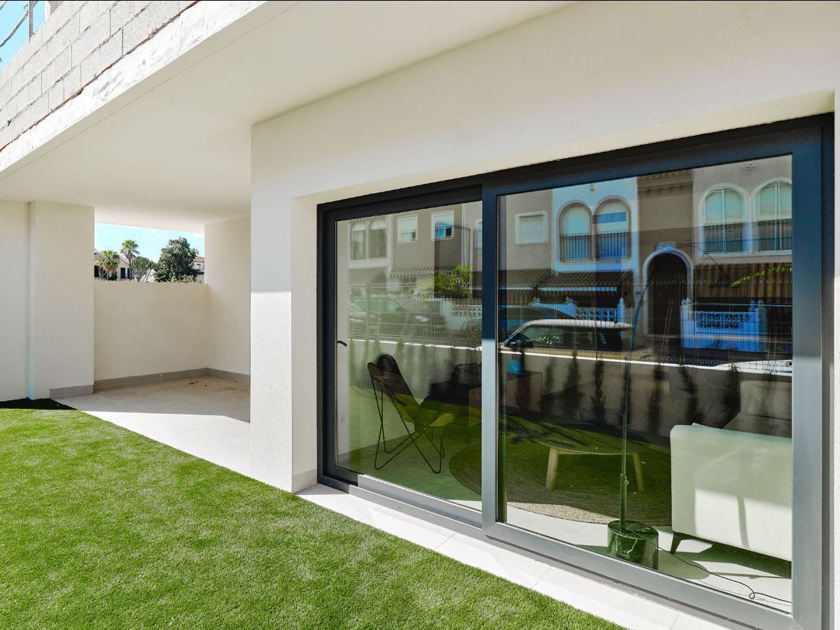 Modern white house exterior with large glass sliding doors, a covered patio, and a green lawn.