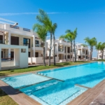 Resort-style apartment complex with a long pool, palm trees, and white modern buildings on a sunny day.