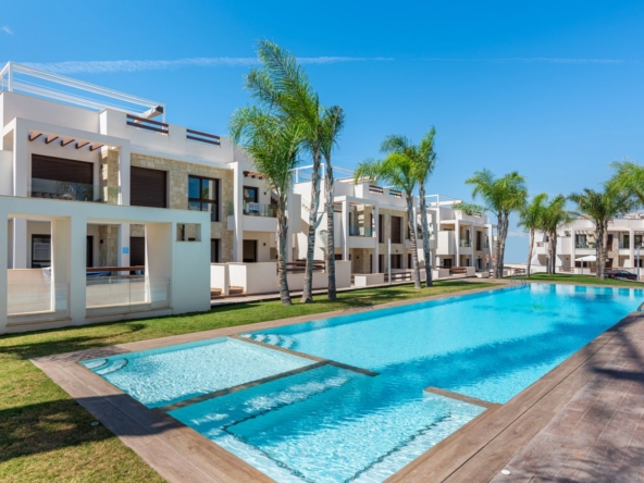 Resort-style apartment complex with a long pool, palm trees, and white modern buildings on a sunny day.
