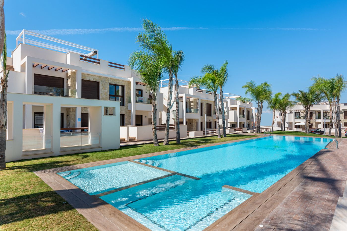 Resort-style apartment complex with a long pool, palm trees, and white modern buildings on a sunny day.