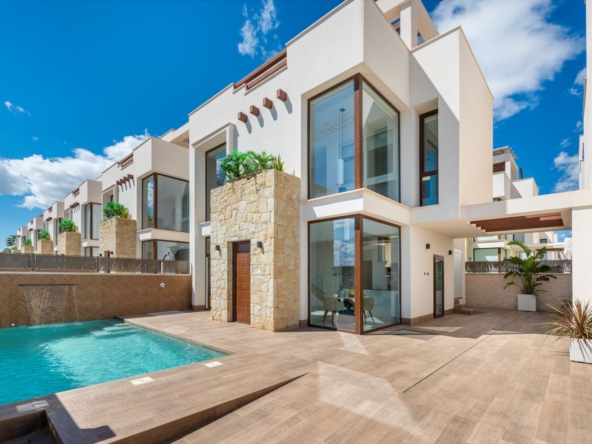Modern white villa with stone accents, large glass windows, and a turquoise pool set on a tiled courtyard under a blue sky.