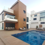 Three-story modern villa with brown brick accent, glass balcony, and a rectangular blue pool in the foreground; outdoor dining and lounge furniture on the patio.