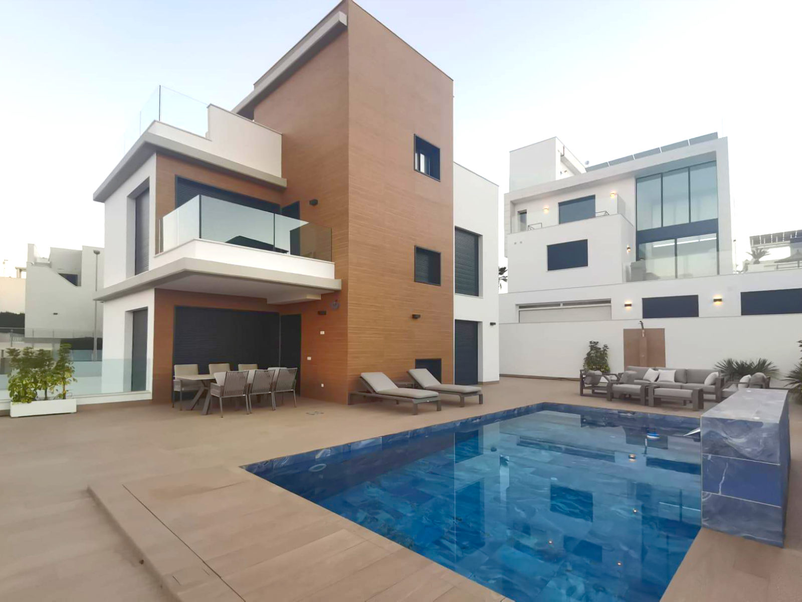 Three-story modern villa with brown brick accent, glass balcony, and a rectangular blue pool in the foreground; outdoor dining and lounge furniture on the patio.