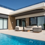 Modern house exterior with stone column, beige seating, and a blue pool in the foreground, under a clear sky.