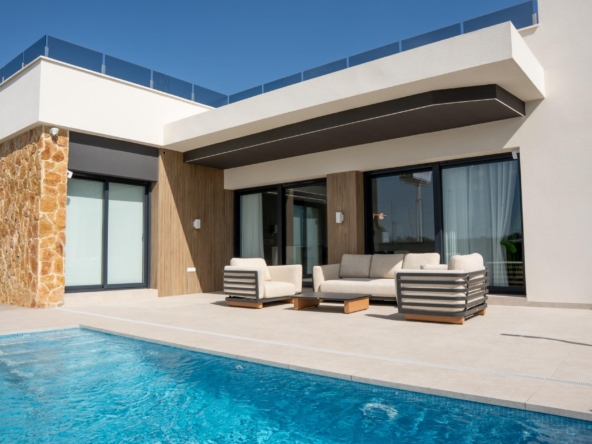 Modern house exterior with stone column, beige seating, and a blue pool in the foreground, under a clear sky.