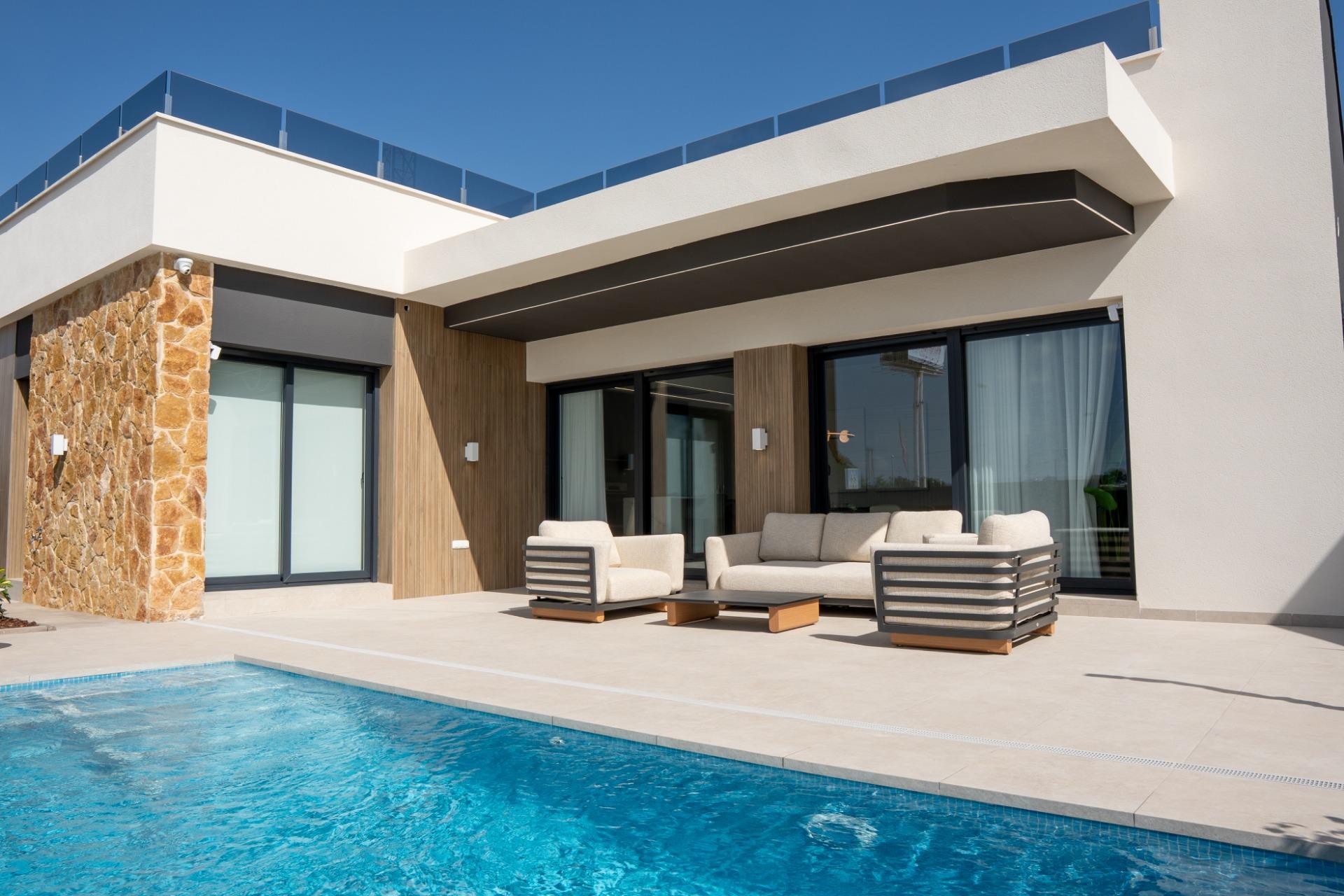 Modern house exterior with stone column, beige seating, and a blue pool in the foreground, under a clear sky.