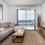 Cozy modern living room with a gray sectional sofa, beige cushions, and a wooden coffee table facing a glass sliding door to a beach view.