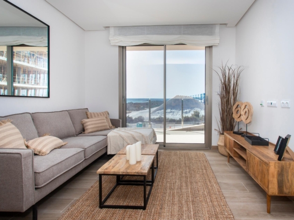 Cozy modern living room with a gray sectional sofa, beige cushions, and a wooden coffee table facing a glass sliding door to a beach view.