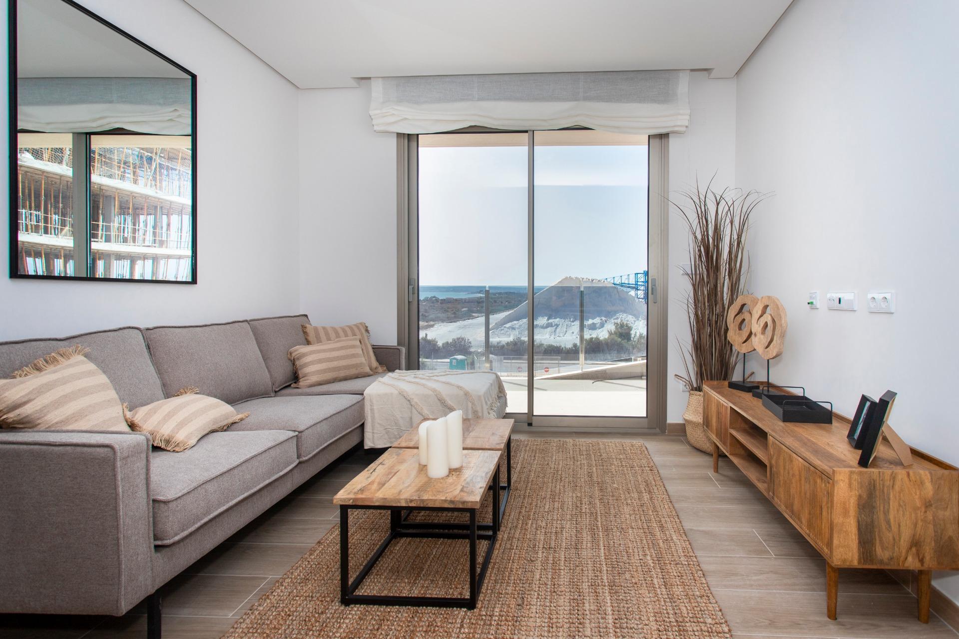 Cozy modern living room with a gray sectional sofa, beige cushions, and a wooden coffee table facing a glass sliding door to a beach view.