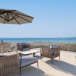 Outdoor seaside terrace with wicker lounge chairs, a large beige umbrella, and glass railing overlooking the ocean.