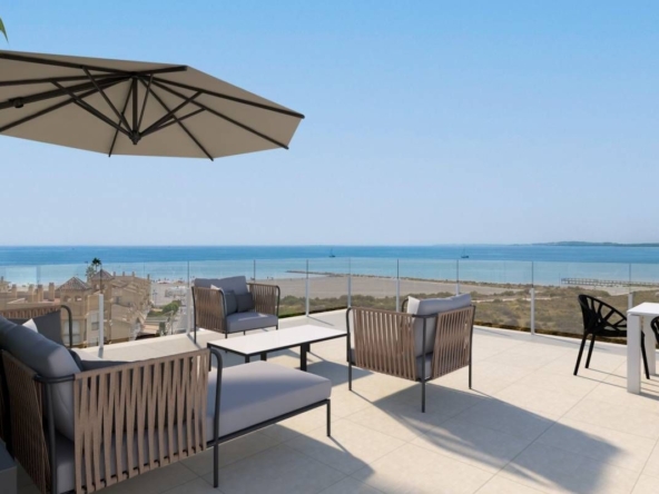 Outdoor seaside terrace with wicker lounge chairs, a large beige umbrella, and glass railing overlooking the ocean.