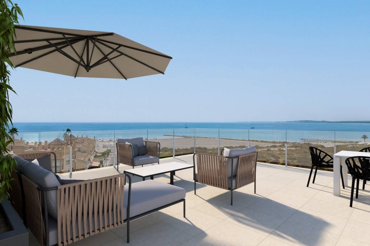 Outdoor seaside terrace with wicker lounge chairs, a large beige umbrella, and glass railing overlooking the ocean.