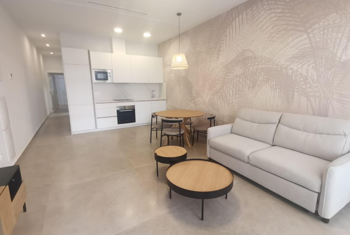 Open-plan living area with a white kitchen, round wooden dining table, and a light gray sofa against a palm-patterned wall, bright and minimalist.