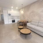Open-plan living area with a white kitchen, round wooden dining table, and a light gray sofa against a palm-patterned wall, bright and minimalist.
