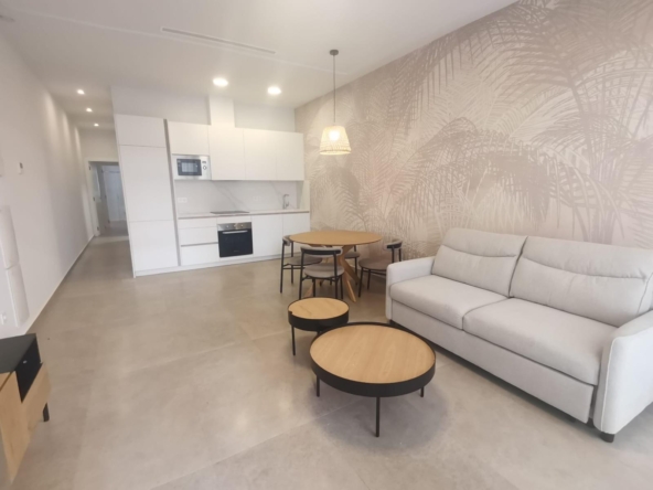 Open-plan living area with a white kitchen, round wooden dining table, and a light gray sofa against a palm-patterned wall, bright and minimalist.