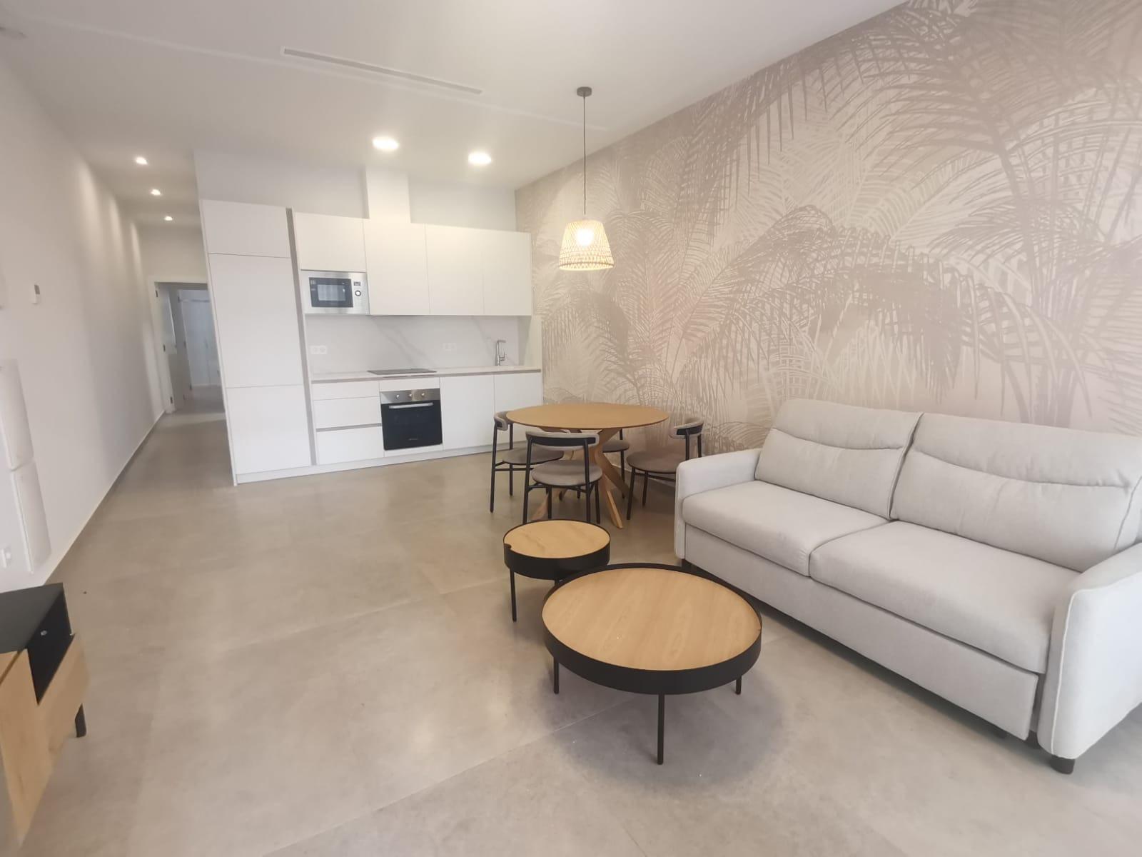 Open-plan living area with a white kitchen, round wooden dining table, and a light gray sofa against a palm-patterned wall, bright and minimalist.