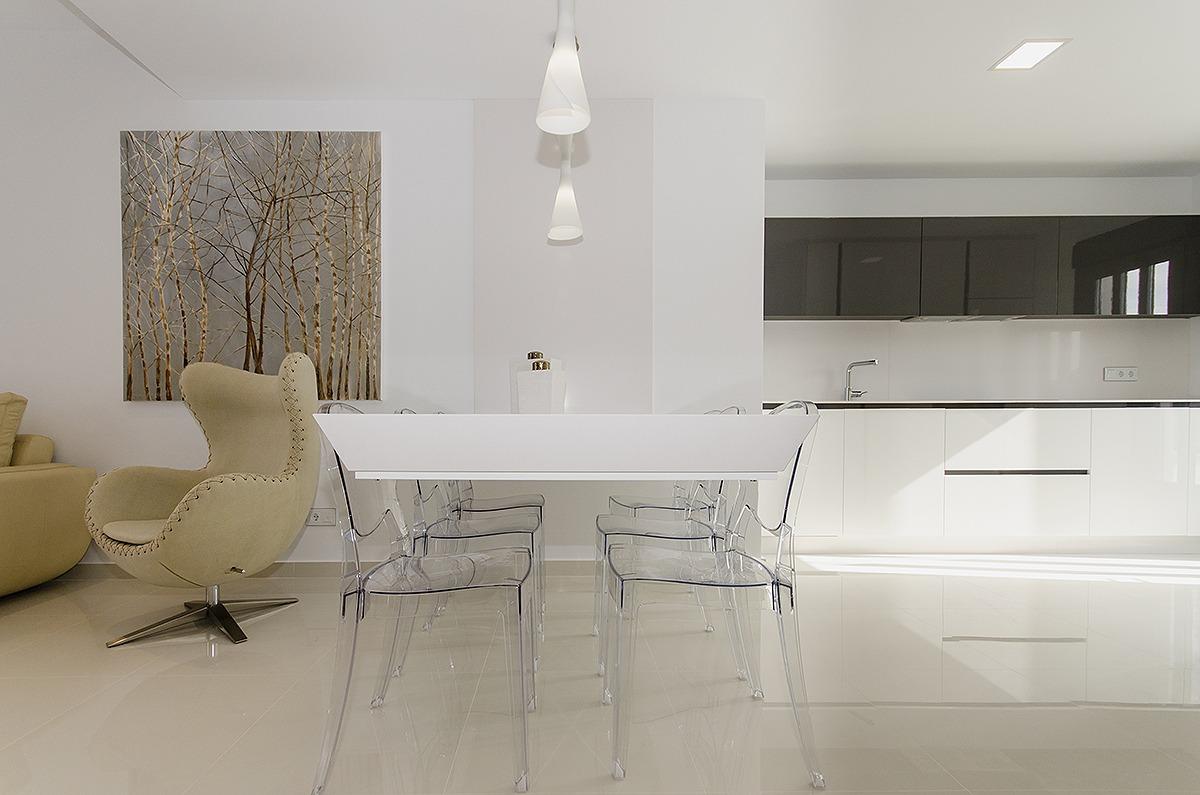 Bright open-plan kitchen and dining area with a white table and clear acrylic chairs; beige curved lounge chair and birch-tree wall art beyond.