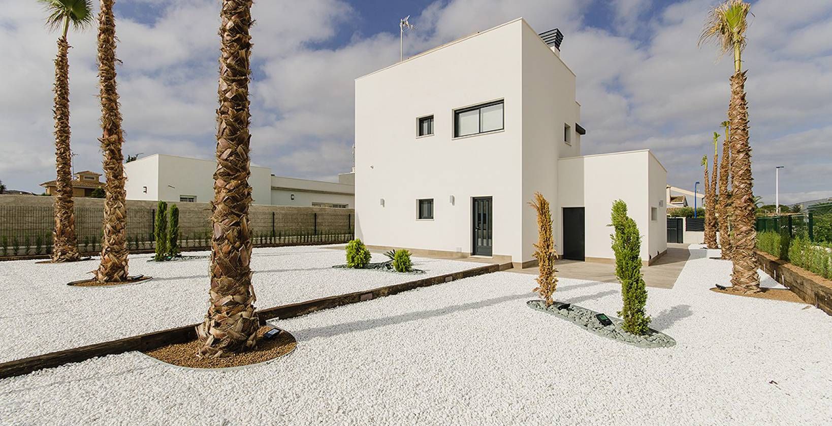 Front view of a modern white two-story house with small windows, set in a palm-tree xeriscape yard with white gravel.