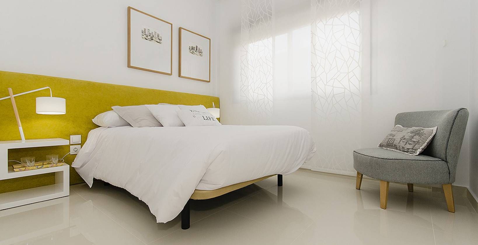 Bright, minimalist hotel-style bedroom with a white bed, yellow headboard, and a gray upholstered chair by a white nightstand.