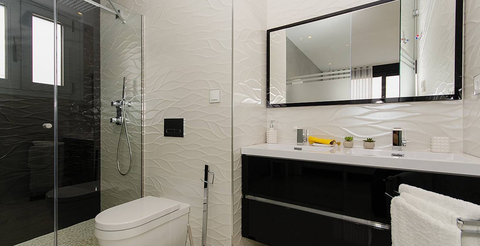 Modern bathroom with a glass-walled shower, wall-mounted toilet, and a white countertop vanity beneath a large framed mirror.