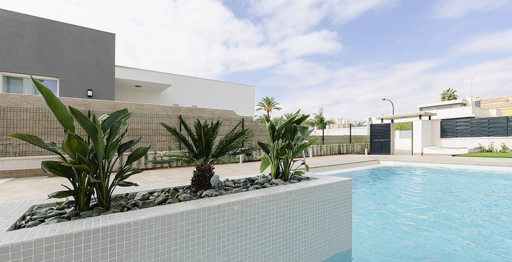 Modern outdoor pool area with a tiled planter box containing palm-like plants and rocks, beige walkway, and white/gray buildings in the background.