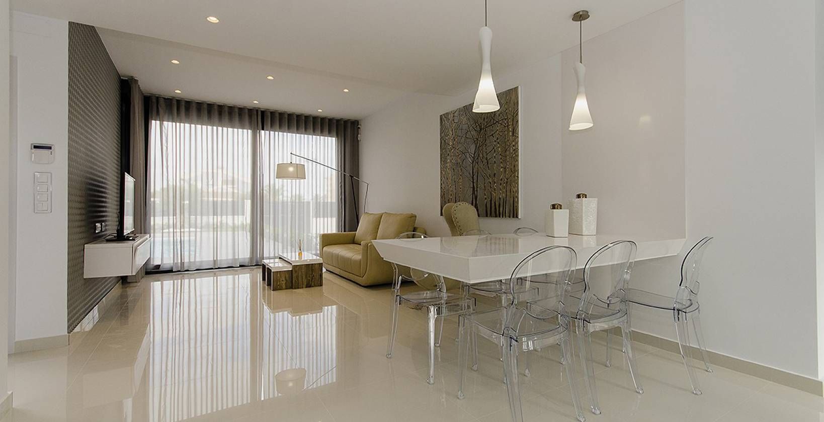 Open-plan living and dining room with a glossy white floor, a white dining table, and clear acrylic chairs; beige sofa and sheer-curtain windows in the background.